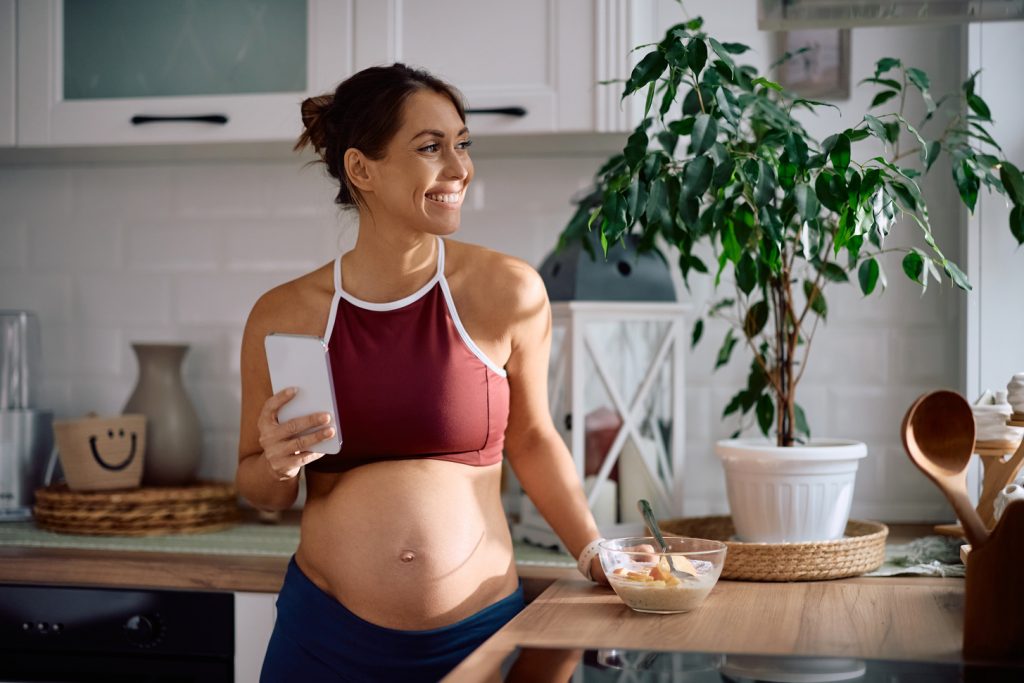 Gestante em casa utilizando celular enquanto se alimenta durante a gravidez.
